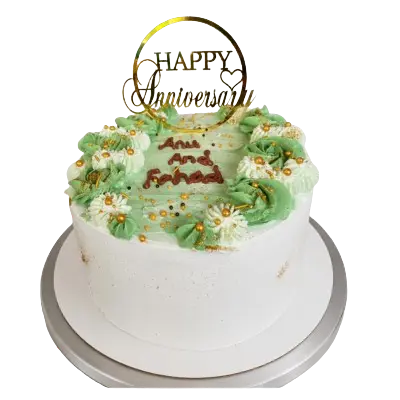 A round white anniversary cake topped with light green frosting, white and green piped flowers, gold sprinkles, and a circular gold 'Happy Anniversary' topper."