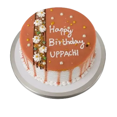 A round birthday cake with a smooth caramel glaze on top and white frosted sides with a caramel drip effect. The top features white frosting stars, gold sprinkles, and white icing text that reads "Happy Birthday UPPACHI".
